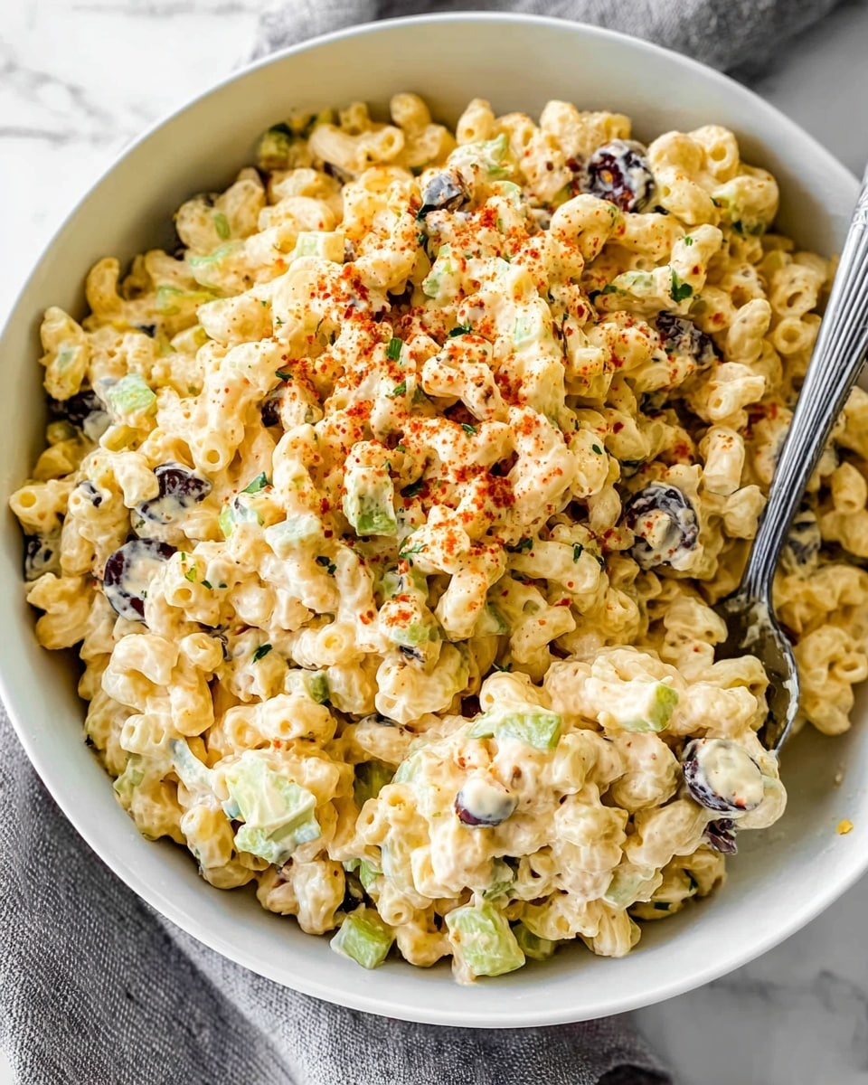 A white bowl filled with creamy macaroni layered throughout with pieces of black olives, small chunks of celery, and finely chopped green herbs. The macaroni is coated in a yellowish dressing, with some light brown specks on the surface, suggesting a sprinkle of seasoning. A silver spoon is scooping a portion from the bowl, revealing the textured, soft pasta mixed with crunchy vegetables and small green bits. The bowl sits on a white marbled surface with soft lighting highlighting the creamy and fresh ingredients. Photo taken with an iphone --ar 4:5 --v 7