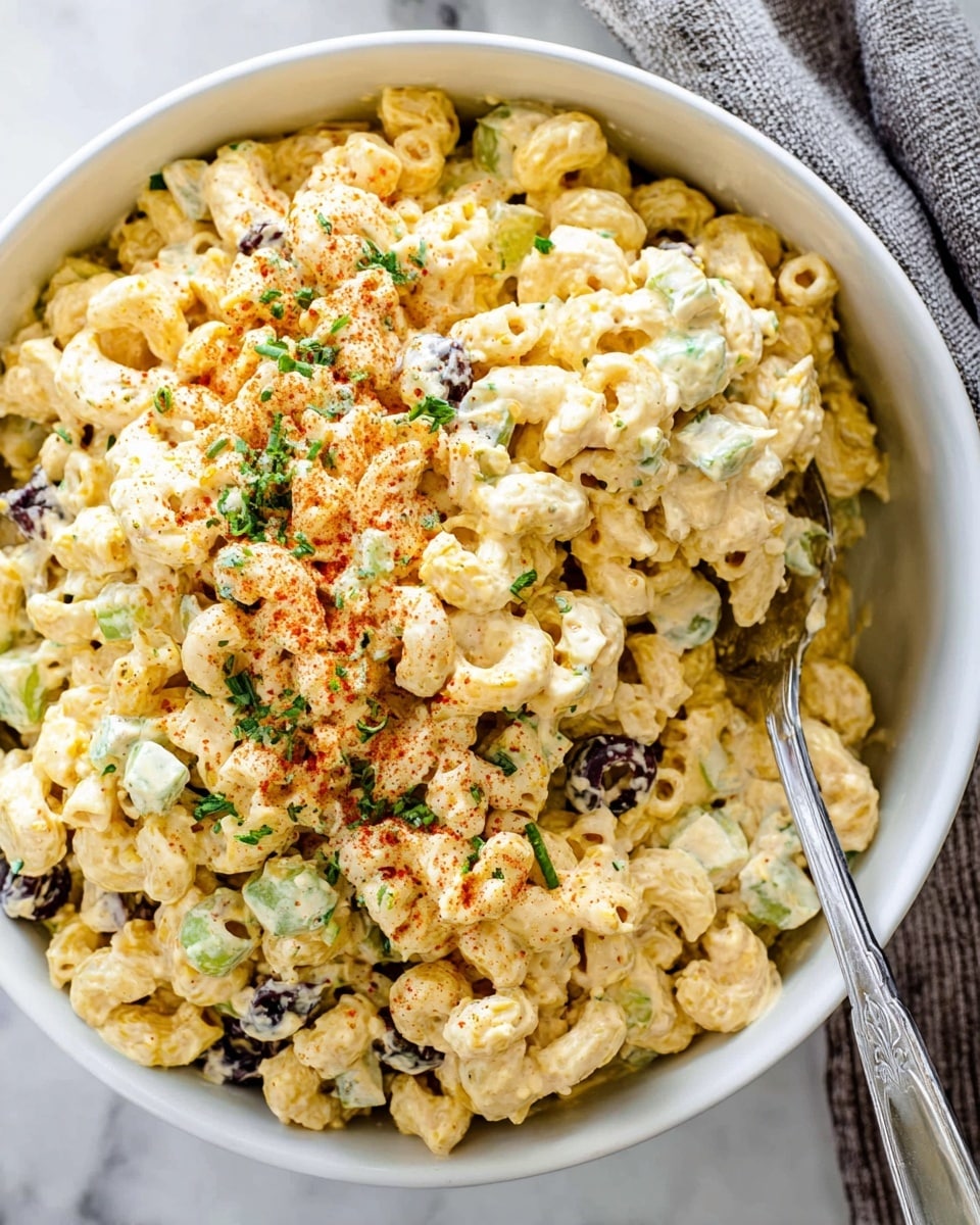 The image shows a white bowl filled with creamy macaroni salad. It has three main visible layers: the top layer has small pieces of green herbs and light red powder sprinkled over pale yellow macaroni mixed with chopped black olives and celery. The middle and bottom layers are dense and creamy macaroni coated in a pale yellow sauce with chunks of celery and bits of black olives scattered throughout. There is a silver spoon inside the bowl scooping some salad. The bowl is placed on a white marbled surface with a gray cloth nearby. Photo taken with an iphone --ar 4:5 --v 7