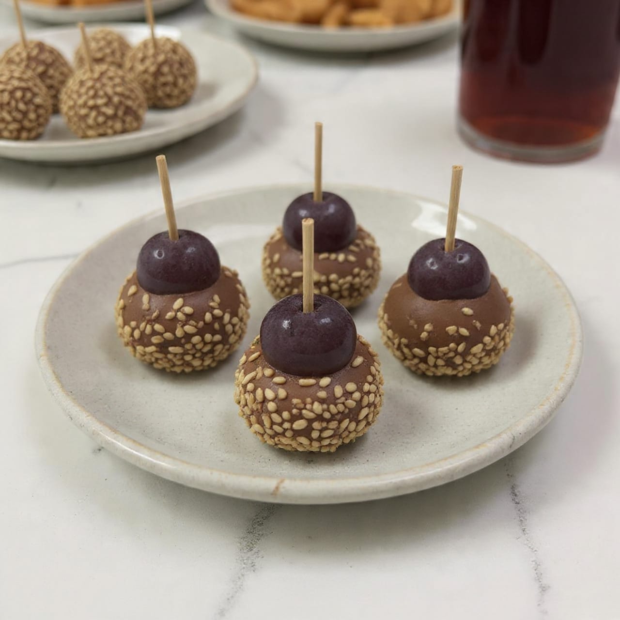 The image shows small round cheese balls coated with a layer of crunchy oats and herbs, placed on a light-colored ceramic plate. Each cheese ball is topped with three dark purple grapes secured with a toothpick standing vertically in the center. The cheese looks creamy white with green herb bits mixed in. The oats stick to the outside, giving a rough texture with brown and golden tones. The plate is set against a white marbled surface and the background is softly blurred with hints of other snacks on a similar light plate and a deep reddish drink. Photo taken with an iphone --ar 4:5 --v 7