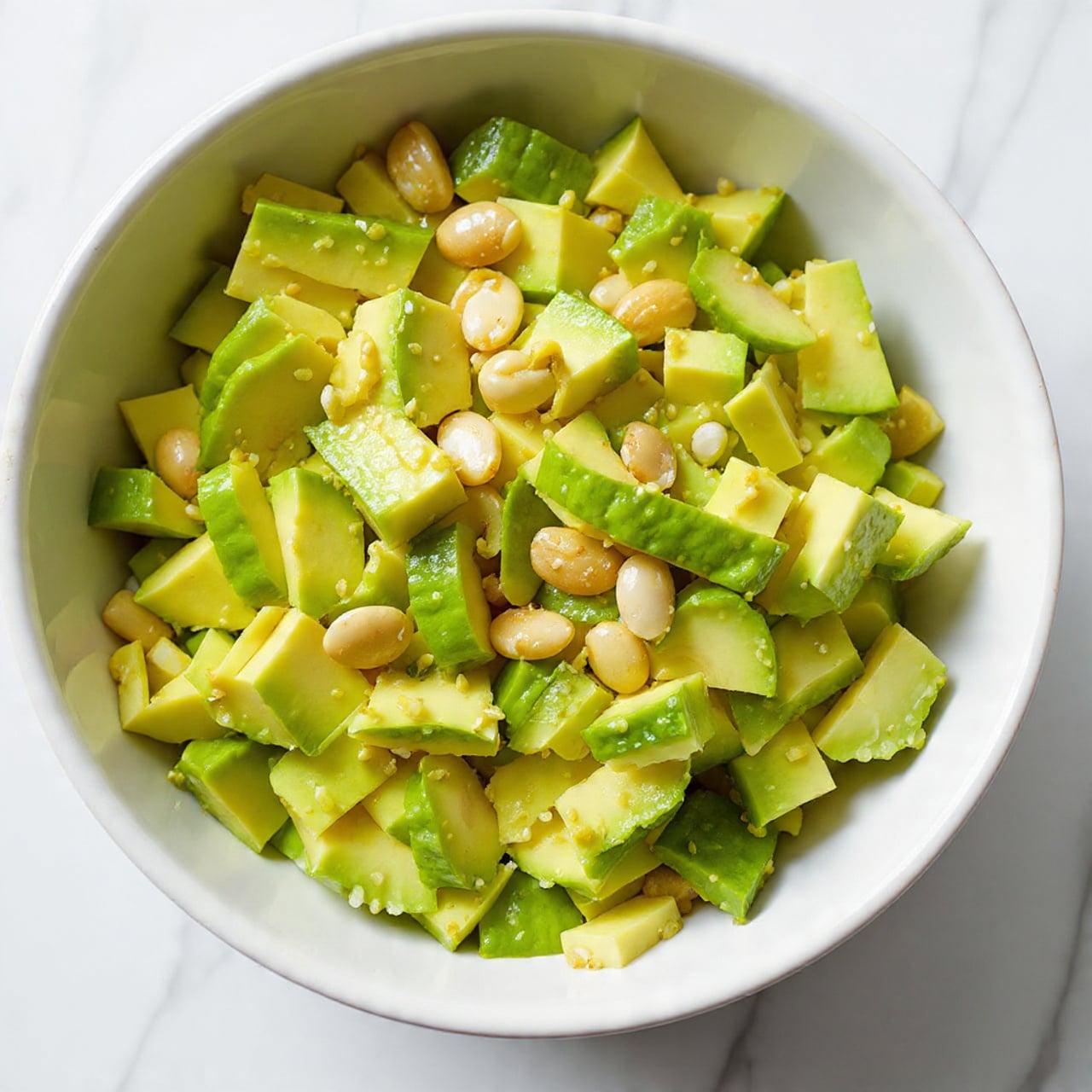 A close-up view of a fresh avocado salad served in a white bowl on a white marbled surface, showing chopped avocado mixed with halved cherry tomatoes, diced cucumber, and thinly sliced red onion, finished with chopped fresh cilantro or parsley and a light sheen from the dressing. Photo taken with an iphone