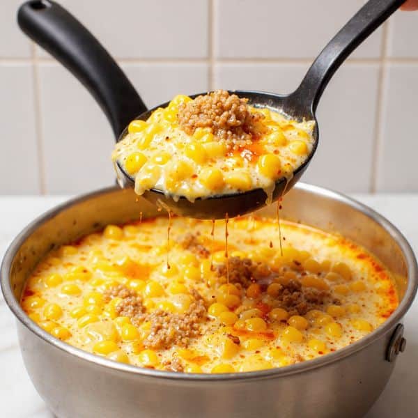 A close-up view shows a large silver pot filled with a creamy soup that has a rich orange tint floating on the surface. A black ladle lifts a scoop of the soup, revealing several layers: at the base, small golden-yellow corn kernels; mixed in are soft tan potato chunks and brownish pieces of ground meat; all coated in a thick, creamy off-white broth with hints of orange oil floating on top. The texture looks smooth but chunky, with the ingredients clearly visible and well mixed. The background features a white marbled surface and a white tiled wall. photo taken with an iphone --ar 4:5 --v 7