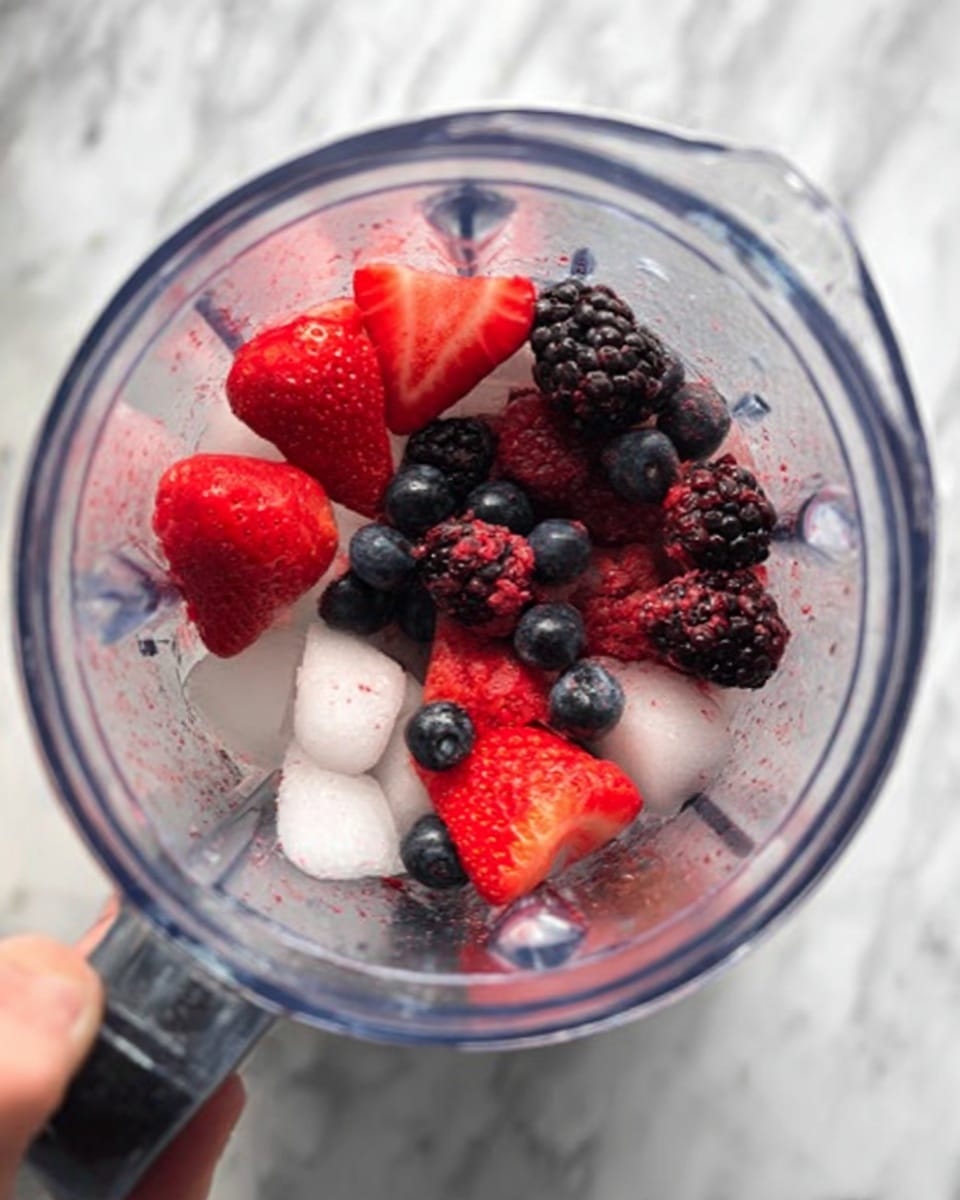 Inside a clear blender container, several bright red strawberry halves and whole blackberries and blueberries sit on top of small white ice cubes at the bottom. The fruit layers show a mix of deep reds, dark purples, and shiny blues, contrasting with the clear and frosty texture of the ice. This whole setup rests on a white marbled surface. A woman's hand is visible holding the blender steady from the side. photo taken with an iphone --ar 4:5 --v 7
