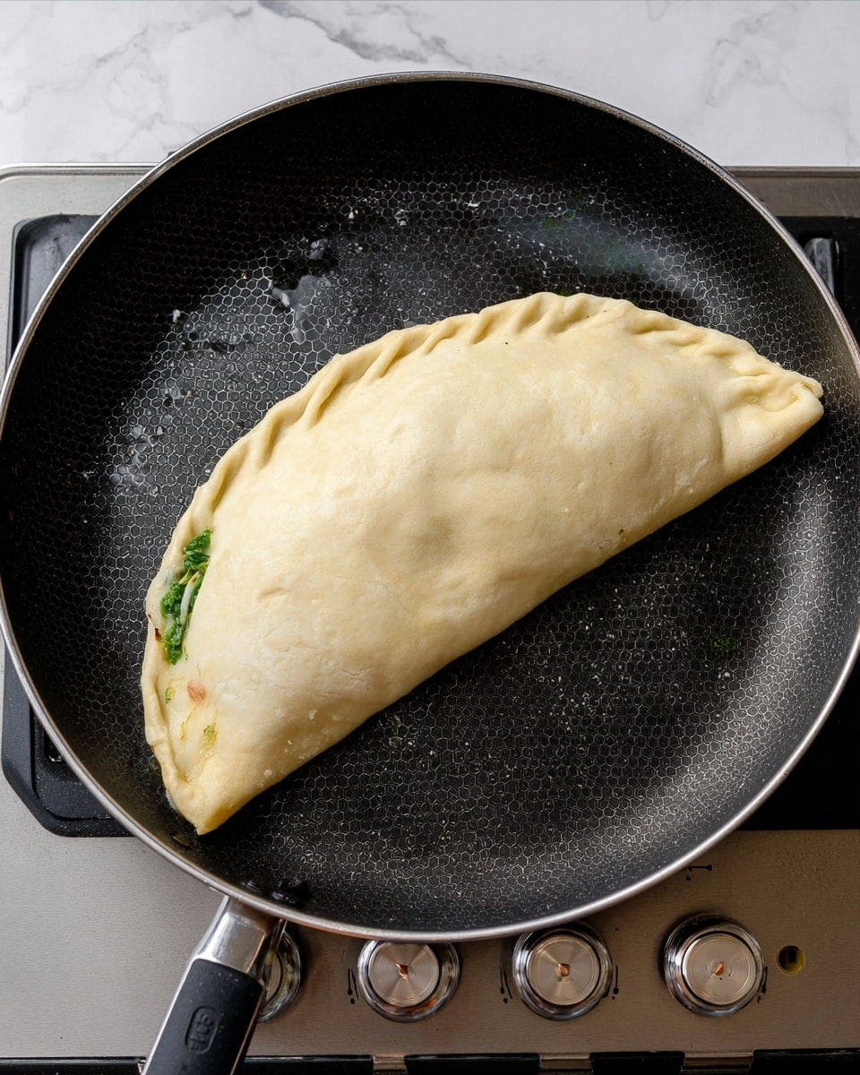 A half-moon shaped calzone with a pale, soft dough exterior is cooking in a black frying pan with small hexagon patterns and droplets of oil scattered around. The edges of the calzone are pressed together, sealing the filling inside, with a slight green peek of spinach or herbs visible near one edge. The frying pan is on a stove with silver and black knobs, and the background shows a white marbled surface. Photo taken with an iphone --ar 4:5 --v 7