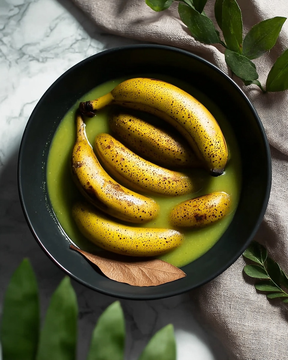 A black bowl holds seven whole bananas with yellow skins that have many small brown spots. The bananas rest in a shallow layer of green sauce with a smooth texture. One piece of dry brown leaf is partly visible near the bottom of the bowl. Around the bowl, there are green leaves and a soft, grey cloth, all placed on a white marbled surface. The lighting casts soft shadows, highlighting the shiny surface of the bananas and the sauce photo taken with an iphone --ar 4:5 --v 7
