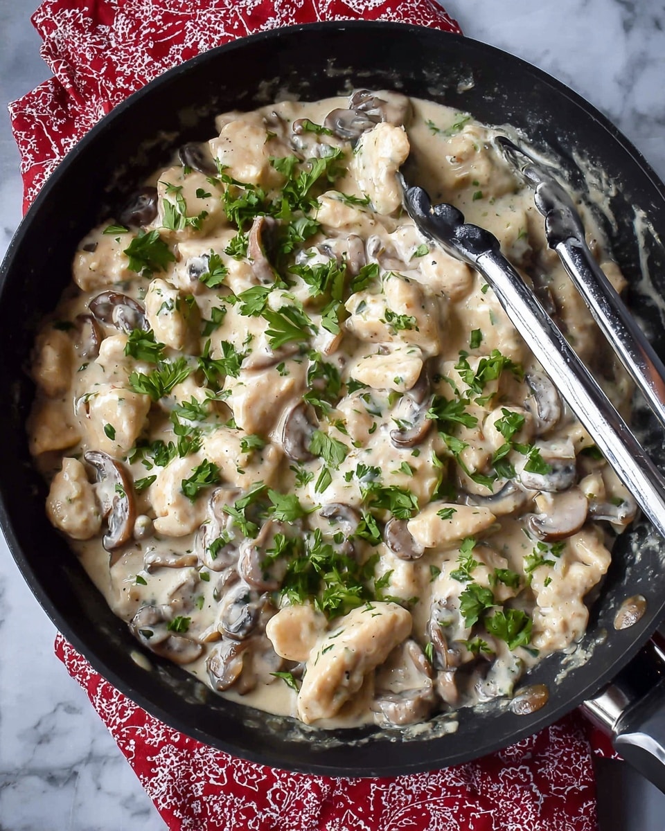 The image shows a black pan full of a creamy dish with several layers. The base layer is made of small white pieces of chicken covered in a thick, shiny white sauce. Mixed in and sitting on this base are dark brown sliced mushrooms giving some texture contrast. Scattered on top are fresh green chopped parsley leaves adding a pop of color. On the right side of the pan, a pair of metal tongs with black handles rests over the dish. The pan sits on a red and white patterned cloth, placed on a white marbled surface. Photo taken with an iphone --ar 4:5 --v 7