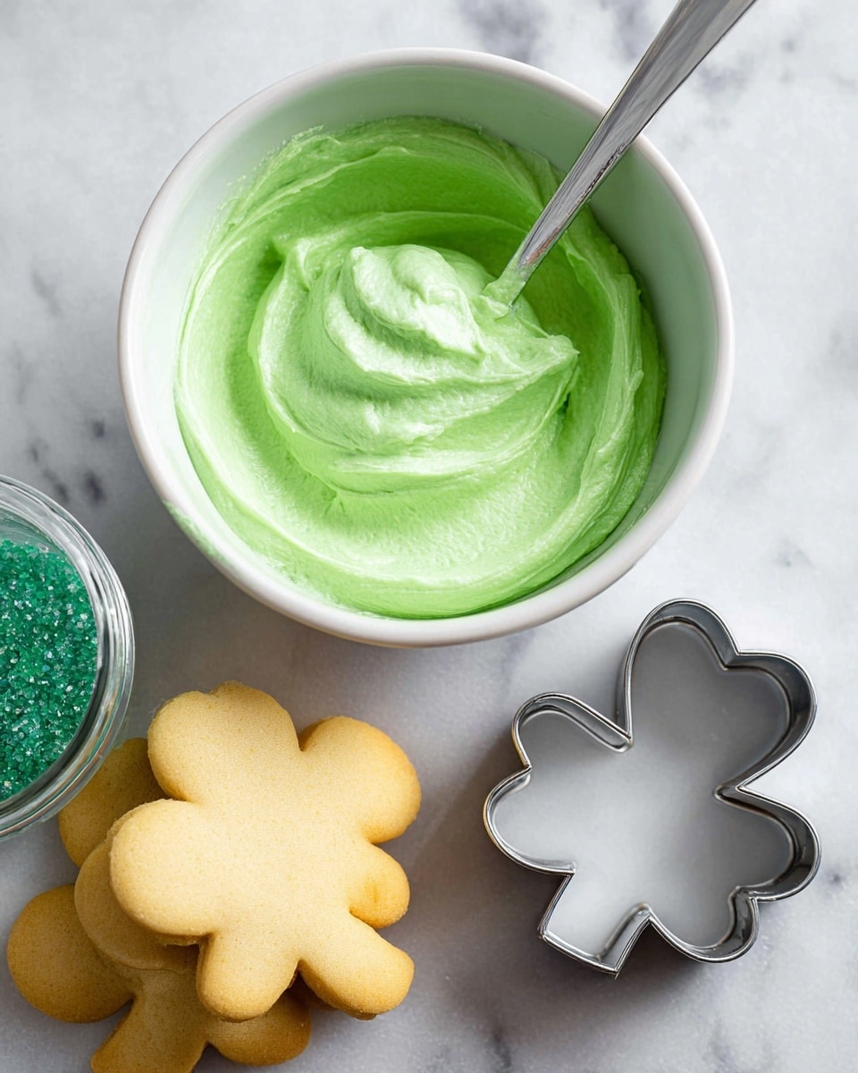 A close-up of a white bowl filled with smooth, bright green frosting, swirled with a spoon inside. Below the bowl, there are two pale golden shamrock-shaped cookies stacked on top of each other. To the right of the cookies is a metal shamrock cookie cutter with a shiny texture. On the left side of the bowl, there is a clear glass jar with a white lid, filled with small, dark green sugar crystals. All items are placed on a white marbled surface. photo taken with an iphone --ar 4:5 --v 7