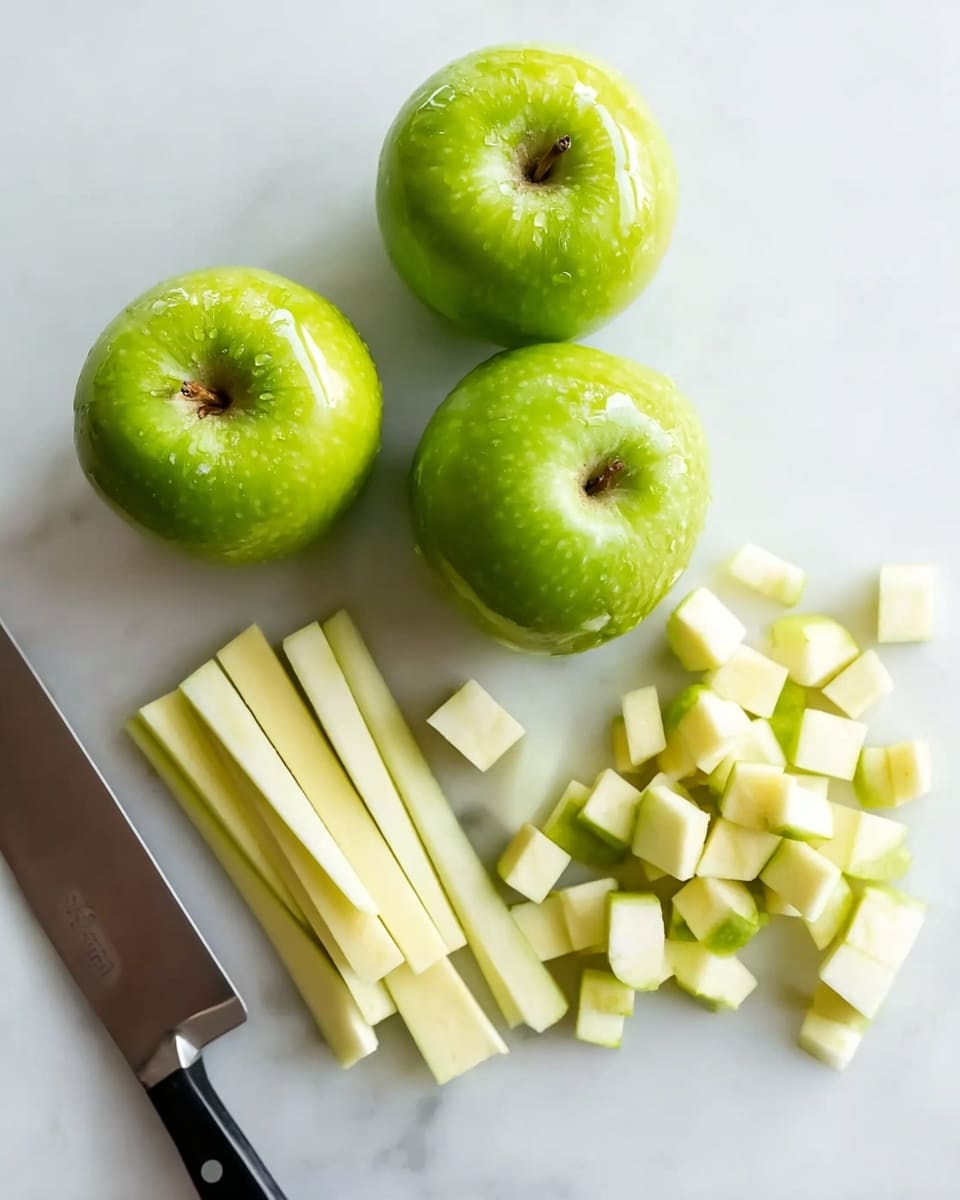 Four whole green apples with shiny skins are placed on the upper part of a white marbled surface. Below them, one apple is sliced into long sticks on the left and small cubes in the middle and right side, showing a pale yellow inside. A large kitchen knife with a dark handle is seen on the bottom left edge, next to the chopped apple pieces. The photo taken with an iphone --ar 4:5 --v 7