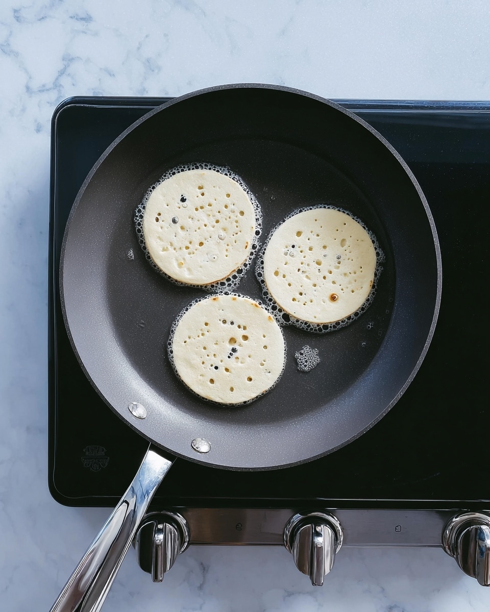 Three small white pancakes cooking in a dark gray pan on a black electric stove. Each pancake has many small bubbles on the surface showing they are cooking. The pan is round with a long silver handle and a curved handle on the opposite side. The background is a white marbled texture. Photo taken with an iphone --ar 4:5 --v 7