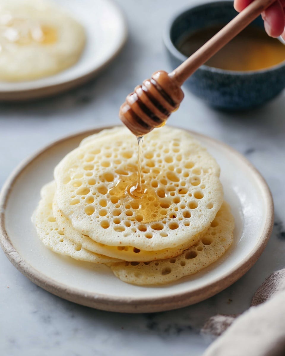 A close-up view of two round, pale golden pancakes with many holes stacked on a white plate, sitting on a white marbled surface. A woman's hand holds a wooden honey dipper above the pancakes, with honey slowly dripping onto the top pancake. In the blurry background, there is a small white plate with another pancake and a dark blue bowl filled with more honey. The overall light is soft and natural, highlighting the texture and the honey's shine. photo taken with an iphone --ar 4:5 --v 7