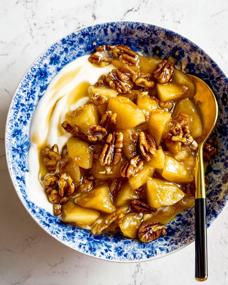 The image shows a white bowl with blue patterns filled with a dessert that has three layers: a smooth white creamy layer at the bottom on the left side, a thick layer of golden brown cooked apple pieces mixed with pecan nuts covering most of the bowl, and a light amber syrup drizzled all over the apple pieces and nuts. A gold spoon with a black handle rests in the bowl among the apple pieces on the right side. The bowl is placed on a white marbled surface. Photo taken with an iphone --ar 4:5 --v 7