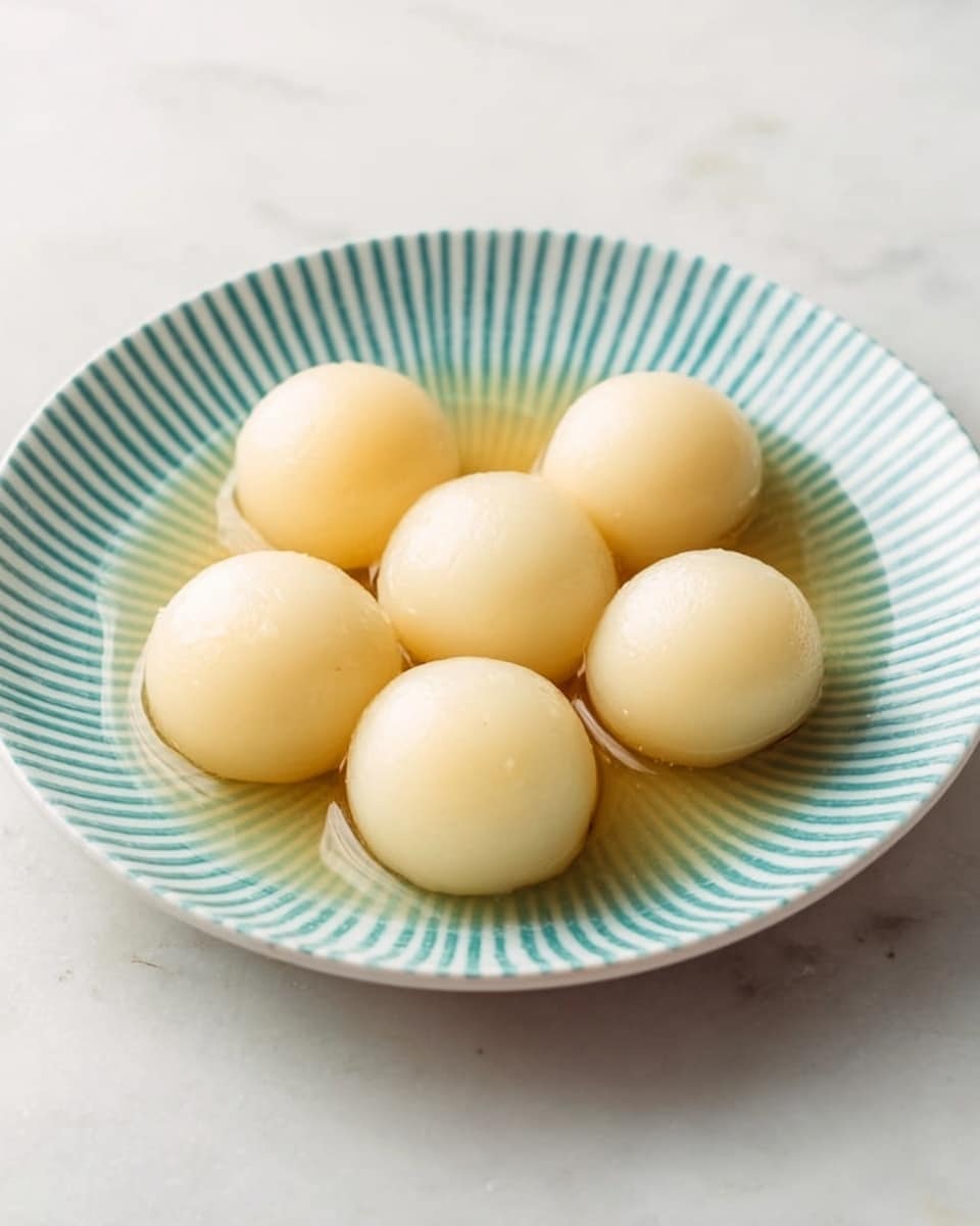 The image shows a white plate with a light blue striped pattern on the rim, holding six smooth, pale yellow balls evenly arranged in two rows of three. The balls have a soft, shiny surface and are sitting in a small pool of clear light brown syrup that covers the plate’s base. The plate is placed on a white marbled surface with soft lighting that highlights the texture and shine of the balls and syrup. Photo taken with an iphone --ar 4:5 --v 7