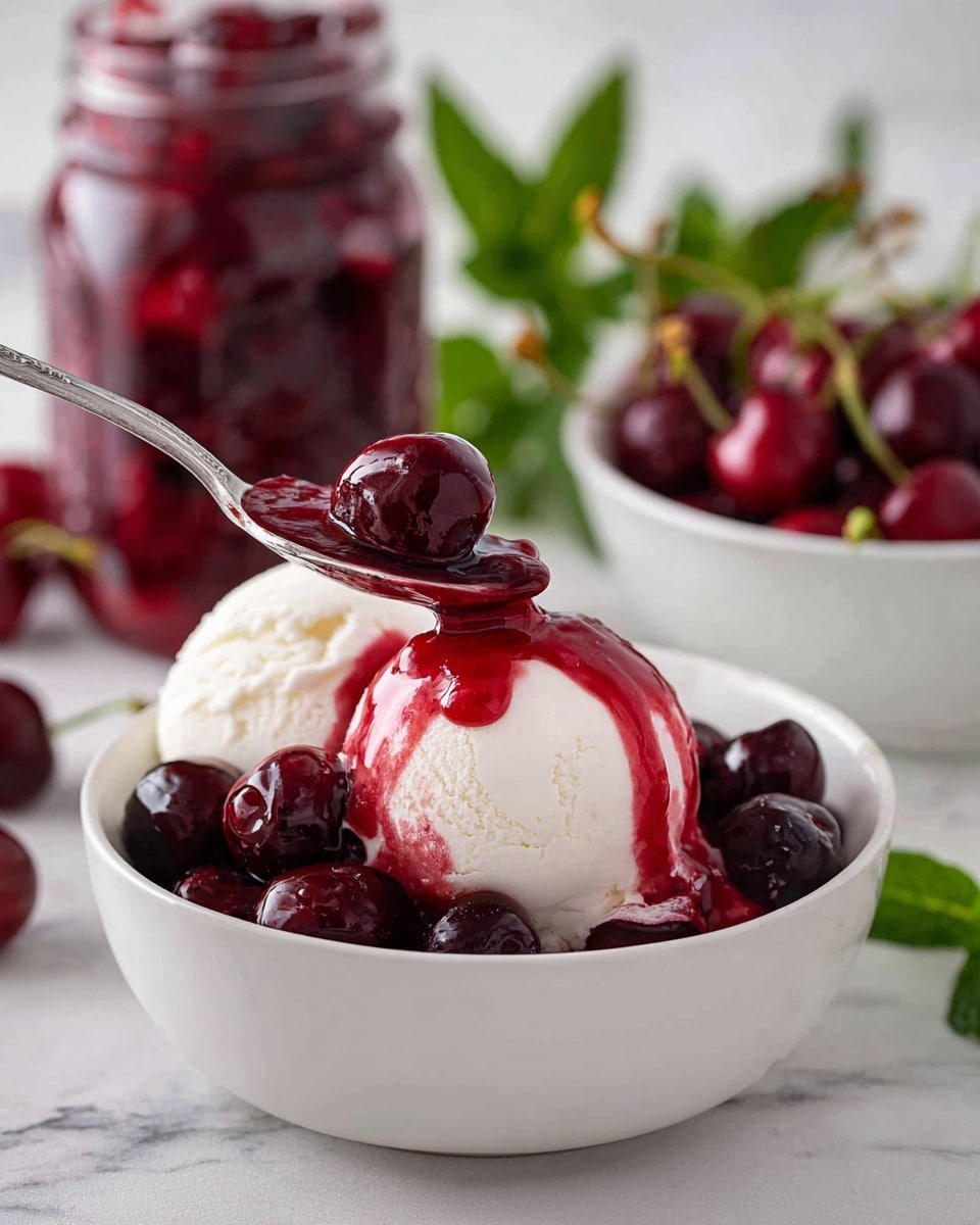 A white bowl filled with two scoops of white ice cream topped with a shiny, deep red cherry sauce that flows down the ice cream’s smooth surface. Dark red whole cherries soaked in the sauce surround the ice cream. A silver spoon lifts a cherry covered in the sauce above the bowl. In the background, a clear glass jar full of cherries in red sauce and a white bowl holding fresh cherries and green mint leaves sit on a white marbled surface with some green leaves visible. Photo taken with an iphone --ar 4:5 --v 7