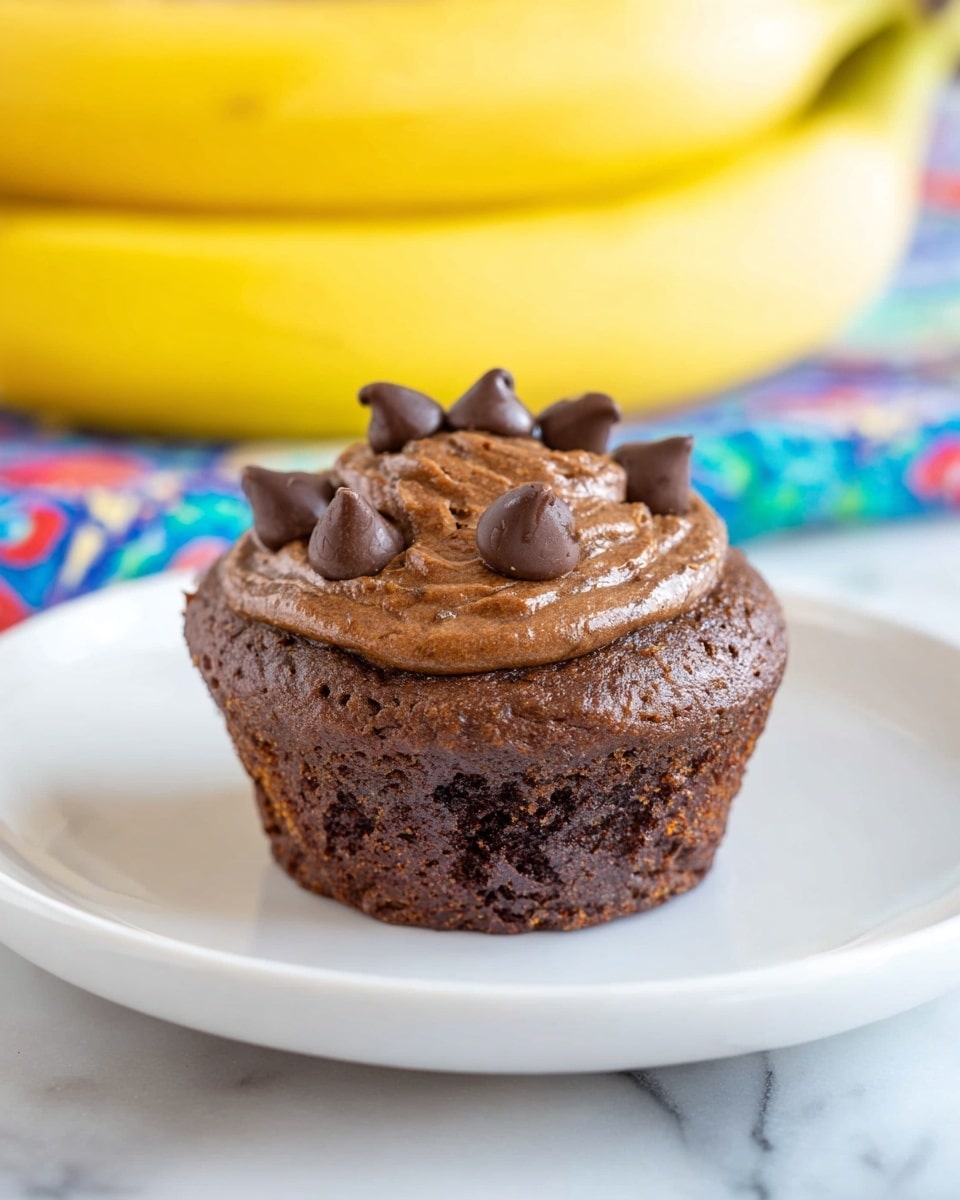 A chocolate muffin sits in the center of a white plate on a white marbled surface. The muffin has two visible layers; the bottom layer is a dense, dark brown base, and the top is a slightly textured, lighter brown domed layer with a smooth swirl on top. There are several chocolate chips scattered on the top of the muffin, adding a glossy contrast. In the background, two bright yellow bananas rest, softly blurred to keep focus on the muffin. A colorful cloth with blue, red, and green patterns peeks from under the plate. photo taken with an iphone --ar 4:5 --v 7