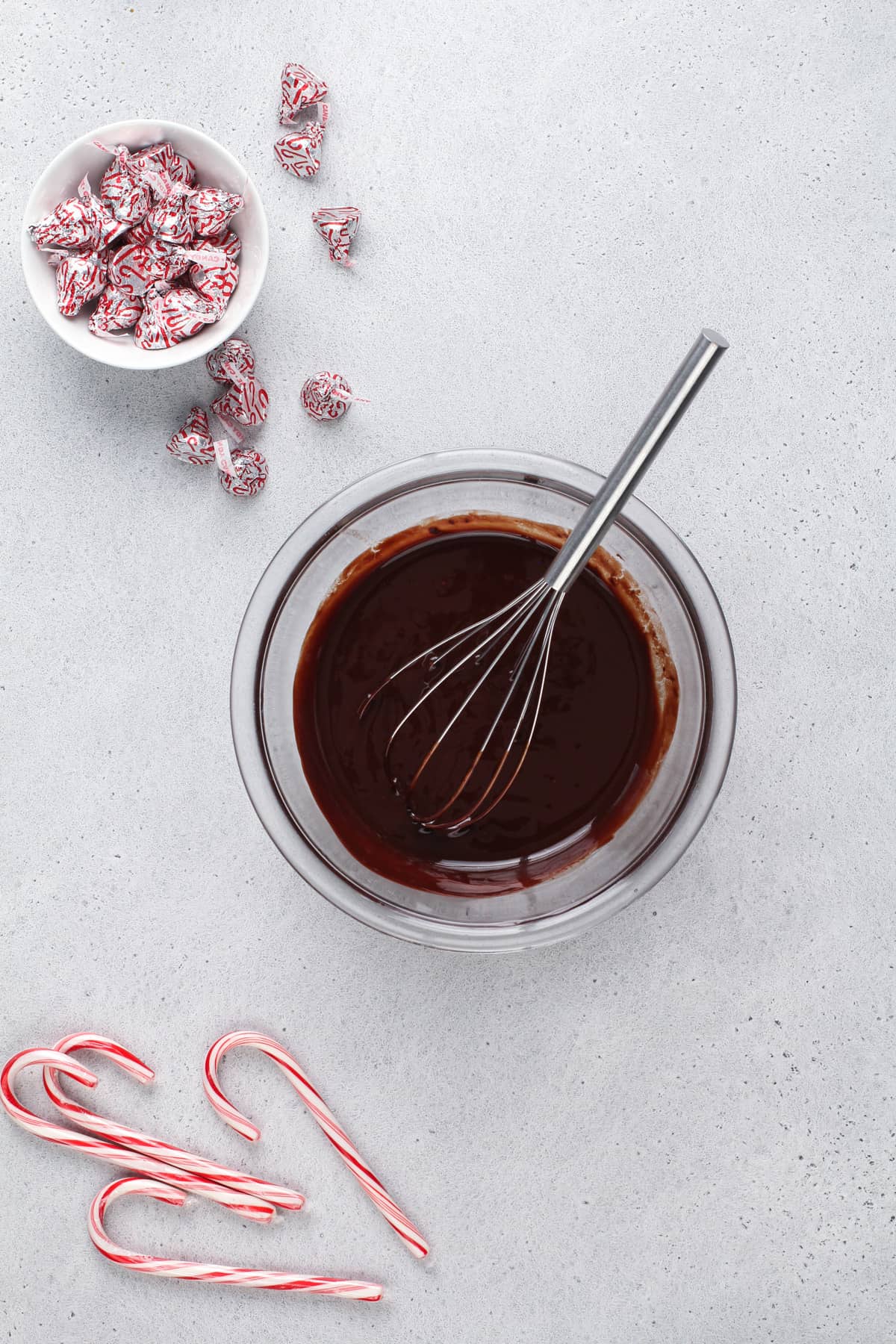 A clear glass bowl filled with a dark, smooth chocolate mixture sits on a white marbled surface, with a stainless steel whisk resting inside the bowl. To the top left, a white bowl contains many small wrapped chocolates in silver foil with red patterns, and some chocolates are scattered nearby on the surface. In the bottom left corner, several red and white striped candy canes are laid out on the white marbled background. photo taken with an iphone --ar 4:5 --v 7