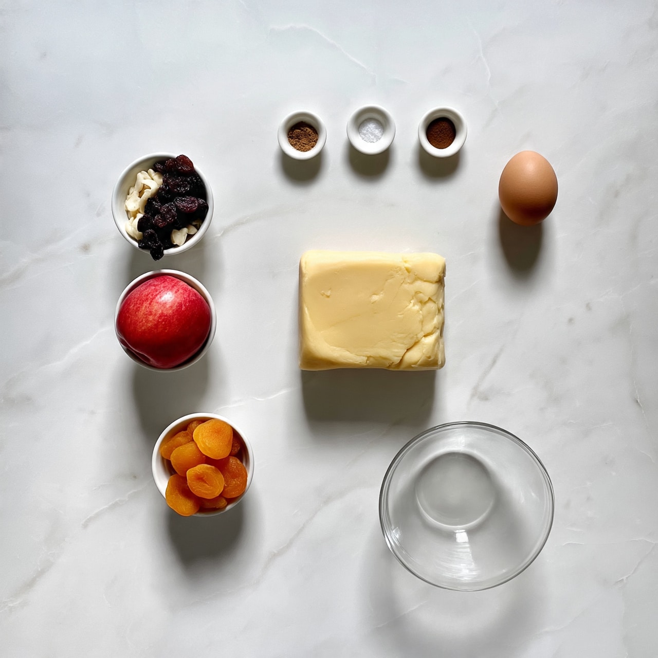 The image shows ingredients laid out neatly on a white marbled surface. In the center, there is a block of light yellow dough or butter, surrounded by two red apples to the left. Around these are small white bowls containing dark raisins, white nuts, and orange dried fruit pieces. A single brown egg is placed on the right side. Near the top, small amounts of brown spices and a pinch of salt are arranged in tiny white bowls. A glass bowl sits empty towards the bottom right. The colors are warm and natural, with a clean and organized layout. Photo taken with an iphone --ar 4:5 --v 7